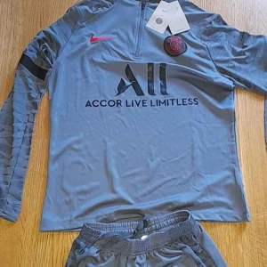 Grå träningsset från Nike - Snyggt grått träningsset från Nike i samarbete  med PSG. Tröjan har långa ärmar och dragkedja vid halsen. Byxorna har elastisk midja och fickor. Perfekt för sport och fritid! Oanvända vid frågor är det bara att skriva