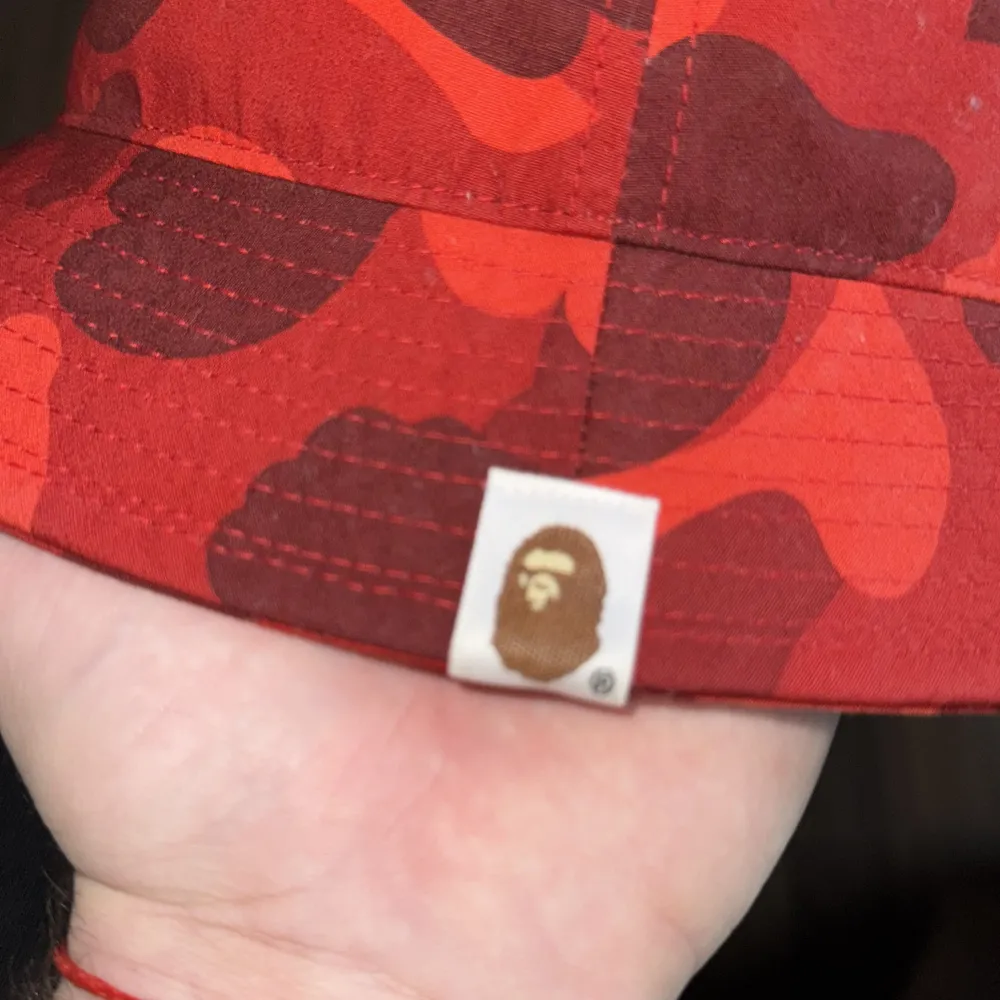 Snygg röd kamouflagehatt från A Bathing Ape med broderad logotyp framtill. Perfekt för att ge din outfit en cool touch. Hatten har en klassisk bucket-modell och är tillverkad i bomull.. Asusteet.