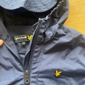 Blå vindjacka från Lyle & Scott - Snygg blå vindjacka från Lyle & Scott med huva och dragkedja. Jackan har ett broderat emblem på bröstet och justerbara dragskor i nederkant. Perfekt för blåsiga dagar.