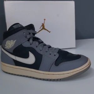 Nike Air Jordan 1 Mid i Grey - Snygga Nike Air Jordan 1 Mid i grått och svart med vita detaljer. Skorna har klassisk snörning och den ikoniska Air Jordan-loggan på sidan. Perfekta för dig som vill ha en stilren och sportig look.