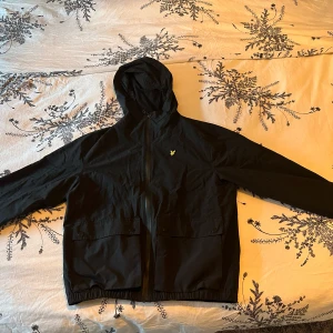 Svart vindjacka från Lyle & Scott - Snygg svart vindjacka från Lyle & Scott med huva och dragkedja. Perfekt för blåsiga dagar. Jackan har en enkel design med en liten logotyp på bröstet och praktiska fickor framtill.