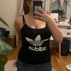 Svart linnetopp från Adidas - Snygg svart linnetopp från Adidas med det klassiska vita logotyptrycket framtill. Perfekt för en sportig och avslappnad stil. Linnet har smala axelband och en bekväm passform.