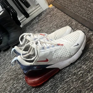 Nike Air Max 270 i vitt och rött - Snygga Nike Air Max 270 sneakers i vitt med röda och blå detaljer. Skorna har en luftig ovandel med snörning och en synlig Air-enhet i hälen för extra komfort. Perfekta för en sportig look.