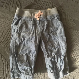 Blå byxor med resår och rosett - Säljer ett par bekväma blå byxor med elastisk midja och en söt rosa rosett framtill. Byxorna har fickor både fram och bak samt resår vid bensluten för en avslappnad look.