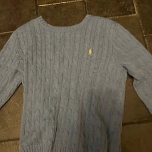 Blå kabelstickad tröja från Ralph Lauren - Säljer en stilren blå kabelstickad tröja från Ralph Lauren med det klassiska logotypbroderiet i gult på bröstet. Tröjan har en rund halsringning och långa ärmar, perfekt för kyligare dagar.