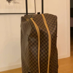 Brun rutig resväska - Snygg brun resväska med klassiskt rutigt mönster och dragkedja. Väskan har ett rymligt inre med mönstrat foder och praktiska hjul för enkel transport.