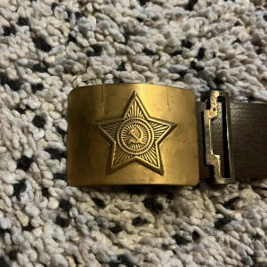 Brunt bälte med stjärnspänne - Snyggt brunt bälte med ett unikt guldfärgat spänne prytt med en stjärna. Perfekt för att ge en cool touch till din outfit. Bältet har en robust design och passar bra till jeans