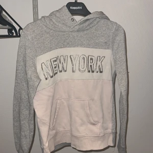 Grå och vit hoodie  - Snygg grå och vit hoodie med texten 'New York' på bröstet. Tröjan har en klassisk känguruficka och en bekväm huva. 