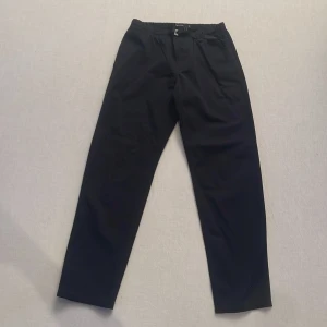 Svarta byxor från Peak Performance - Snygga svarta byxor från Peak Performance med elastisk midja och justerbart bälte. Perfekta för en stilren look med praktiska fickor baktill. Brukar använda de till golf men funkar vardagligt också. Strl 31/34