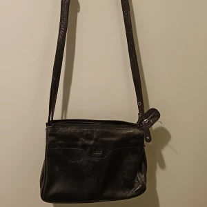 Svart axelväska från Casal - Snygg svart axelväska i skinn från Casal. Väskan har en justerbar axelrem och flera praktiska fack med dragkedja för enkel organisering. Perfekt för att bära med sig det viktigaste på ett stilfullt sätt. Säljer den här väskan av märket Casal. Använd en del, se skick på bilderna. Jag kan skicka fler bilder. Det är mest insidan som har blivit sliten, men funktionen finns. Axelband är justerbart.  26x19 Undersidan 9cm djup. 