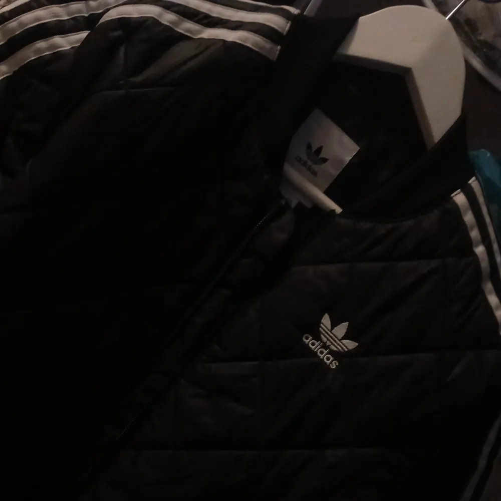 Snygg svart quiltad bomberjacka från Adidas med de klassiska tre ränderna längs ärmarna. Jackan har dragkedja framtill och en sportig look. Perfekt för en casual stil.. Takit.