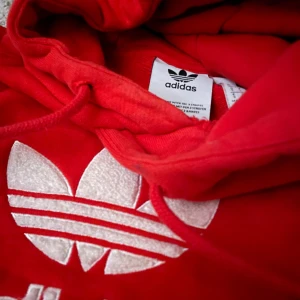 Röd hoodie från Adidas - Säljer en snygg röd hoodie från Adidas med det klassiska trefoil-logotypen i vitt på bröstet. Hoodien har en justerbar huva med dragsko och en stor magficka framtill. Perfekt för en sportig och avslappnad stil.