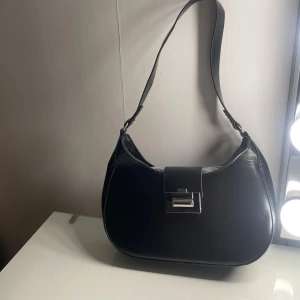 Svart axelväska med metallspänne - Snygg svart axelväska i skinn med ett stilrent metallspänne framtill. Väskan har en enkel och elegant design, perfekt för att komplettera din outfit. Ej använd 