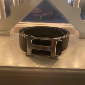 Bälte  - Snyggt svart skärp med ett stort, silverfärgat H-format spänne. Perfekt för att ge en stilren touch till din outfit. Skärpet är tillverkat i skinn och har en elegant design.