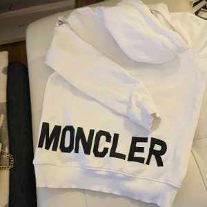 Vit hoodie från Moncler - Säljer en stilren vit hoodie från Moncler med stor logga på framsidan och broderad logotyp på ärmen. Tröjan har en klassisk design med huva och ribbade muddar. Nypris 2200