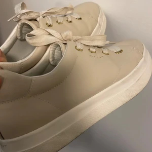 Beige sneakers från Gant - Stilrena beige sneakers från Gant med snörning och en tjock vit sula. Skorna har en diskret logga på sidan och är tillverkade i syntetmaterial. Perfekta för en avslappnad look. Köpta för 1500kr. 