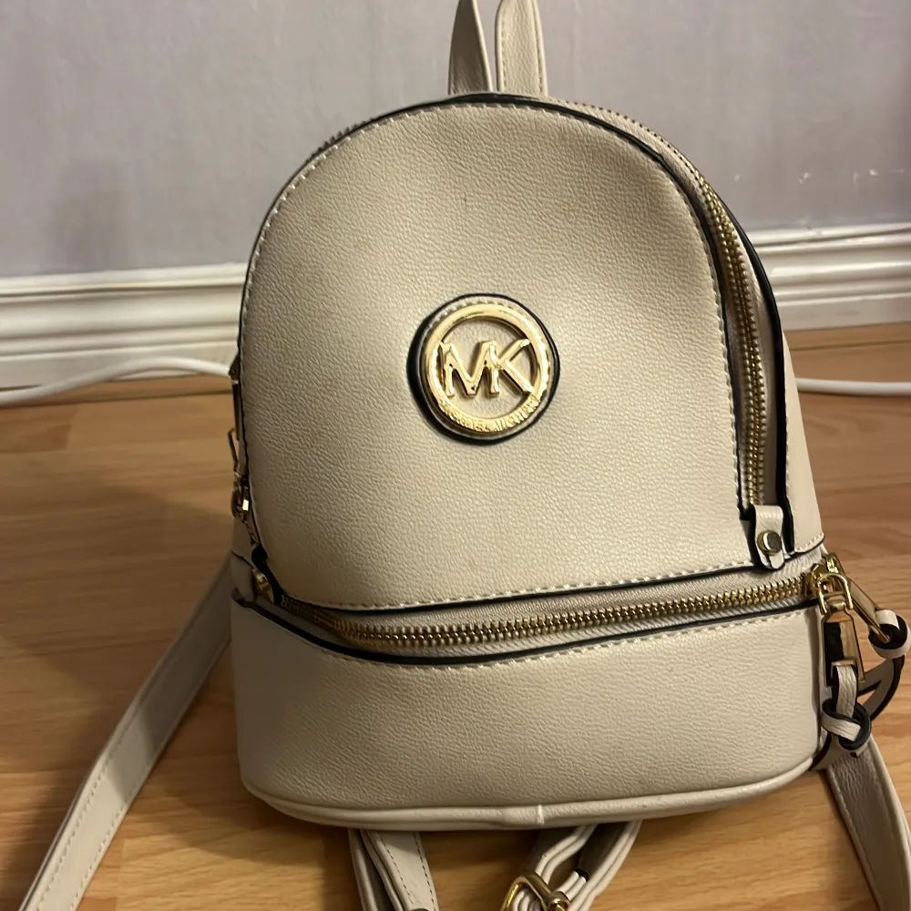 Snygg beige ryggsäck från Michael Kors med guldfärgade detaljer och dragkedjor. Perfekt för en stilren look med det ikoniska MK-emblemet på framsidan. Tillverkad i syntetmaterial och har justerbara axelremmar för bekväm passform.. Laukut & Käsilaukut.