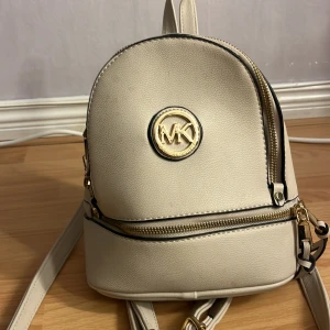 Beige ryggsäck från Michael Kors - Snygg beige ryggsäck från Michael Kors med guldfärgade detaljer och dragkedjor. Perfekt för en stilren look med det ikoniska MK-emblemet på framsidan. Tillverkad i syntetmaterial och har justerbara axelremmar för bekväm passform.