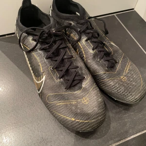 Svarta fotbollsskor från Nike - Säljer ett par svarta fotbollsskor från Nike med snygga gulddetaljer och snörning. Skorna har en slitstark sula med dobbar för bra grepp på planen. Perfekta för fotbollsträning och matcher. original säljs de för 3200 men jag säljer dem för 1000. Väldigt bra skick!