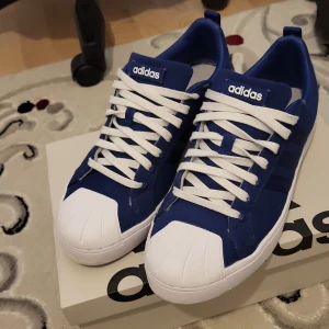 Blå och vita sneakers från Adidas - Snygga blå sneakers från Adidas med vita detaljer och snörning. Perfekta för en sportig look med klassisk design.