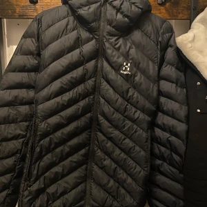 Svart dunjacka från Haglöfs - Snygg och funktionell svart dunjacka från Haglöfs med quiltad design. Jackan har en praktisk huva och dragkedja framtill, perfekt för kyliga dagar. Den är lätt och smidig att bära, vilket gör den idealisk för både stad och natur.