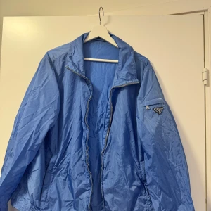Blå vindjacka från Prada - Snygg blå vindjacka från Prada i 100% nylon med resinbeläggning. Jackan har en dragkedja framtill och en klassisk krage. Perfekt för blåsiga dagar och ger en stilren look. Använd fåtal gånger. Jackan har legat i garderoben en längre tid nu så lägger upp den för försäljning. 