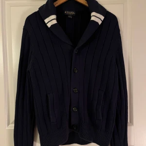 Mörkblå kofta från Brooks Brothers - Säljer en stilren mörkblå kofta från Brooks Brothers i 100% bomull. Koftan har ribbade detaljer och knappar framtill samt en krage med vita ränder. Perfekt för en klassisk look.