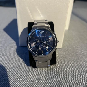 Silverfärgad klocka från Emporio Armani - Snygg silverfärgad klocka från Emporio Armani med blå urtavla och flera sub-dials. Klockan har en stilren design med metallarmband och tydliga visare. Perfekt för den som vill ha en elegant accessoar.