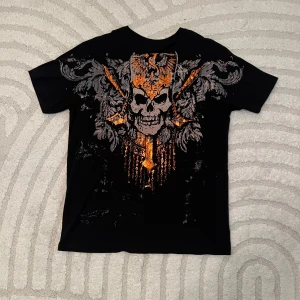 Svart t-shirt med dödskalletryck affliction ish - Cool svart t-shirt med ett stort orange och grått dödskalletryck på framsidan och ett vinge- och ögonmotiv på baksidan. Perfekt för en edgy stil.
