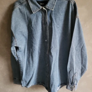 Blå jeansskjorta från Bik Bok - Snygg blå jeansskjorta från Bik Bok i storlek XS. Skjortan har långa ärmar och knappar framtill. Perfekt som jacka för en avslappnad stil med en touch av denim.