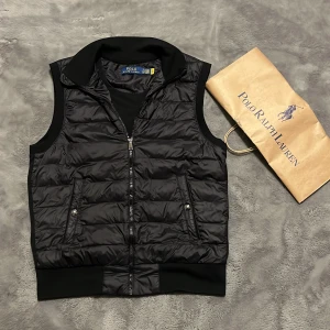 Svart dunväst från Polo Ralph Lauren - Säljer en stilren svart dunväst från Polo Ralph Lauren.Perfekt för lager-på-lager under kyliga dagar. Nästan aldrig använd. Ny pris ca 3000kr! Påse följer med. Öppen för byten !😁