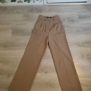 Beige kostymbyxor från Bik Bok - Snygga beige kostymbyxor från Bik Bok med hög midja och vida ben. Perfekta för en stilren look. Byxorna har hällor för bälte och veckade detaljer framtill.