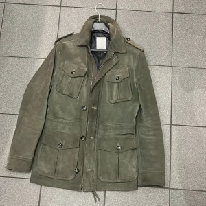 Grön mockajacka från J.Lindeberg - Snygg grön mockajacka från J.Lindeberg med knappar och dragkedja. Jackan har flera praktiska fickor och axelklaffar för en stilren look. Perfekt för höst och vår. Ny pris 11,499kr