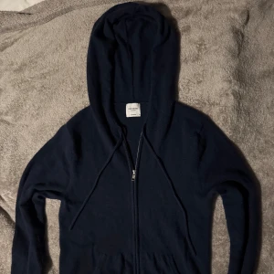 John Henric cashmere hoodie - Säljer en stilren mörkblå hoodie från John Henric med dragkedja och praktiska fickor framtill. Perfekt för kyliga dagar med sin mjuka och bekväma passform. nypris, 2500kr.
