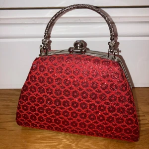 Röd glittrig handväska med mönster - Snygg röd handväska med glittrigt mönster och ett elegant handtag i metall. Väskan har en klassisk stängning upptill och en rymlig insida med fack för enkel organisering.