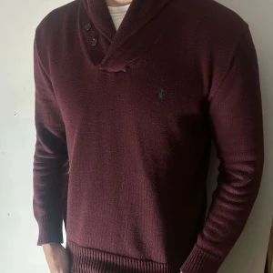 Vinröd tröja från Polo Ralph Lauren - Säljer en stilren vinröd tröja från Polo Ralph Lauren med knappar vid kragen. Tröjan har långa ärmar och är perfekt för kyligare dagar. Den klassiska loggan är broderad på bröstet.