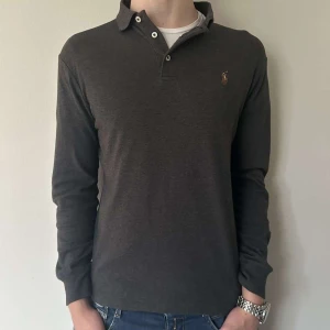 Mörkgrå pikétröja från Ralph Lauren - Snygg mörkgrå pikétröja från Ralph Lauren med klassisk krage och knappar. Tröjan har ett broderat logotypmärke på bröstet och långa ärmar. Perfekt för en stilren look.
