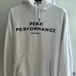 Vit hoodie från Peak Performance - Säljer en snygg och bekväm vit hoodie från Peak Performance. Den har en klassisk design med svart text på bröstet och en praktisk magficka. Lite slitage vid armmuddarna annars i bra skick (ses sista bild). Köpt för ungefär 1200 kronor.