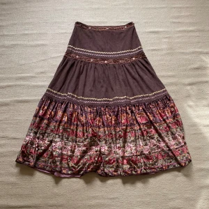 Brun långkjol från Jaune Rouge - Säljer en superfin brun långkjol från Jaune Rouge. Kjolen har ett härligt mönster i varma färger som röd och grön, och är dekorerad med söta broderade detaljer. Perfekt för höst och vår, och funkar lika bra till vardags som till fest. Den är i bra skick och redo för nya äventyr!