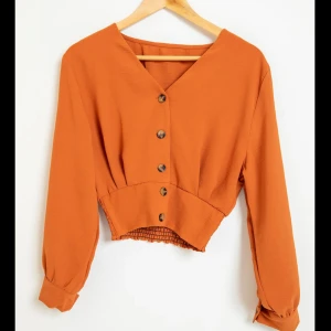 Orange långärmad topp med knappar - Säljer en superfin orange topp med långa ärmar och knappar framtill. Den har en v-ringad design och en elastisk midja som ger en snygg passform. Perfekt för höst och vår!