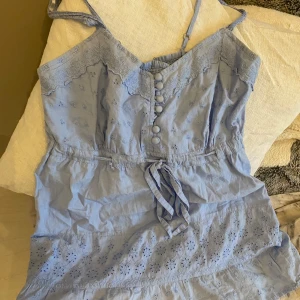 Ljusblå topp med spetsdetaljer - Säljer en superfin ljusblå topp med spetsdetaljer och knappar framtill. Den har smala axelband och ett knytband i midjan för en perfekt passform. Toppen är dekorerad med ett sött blommönster och har en romantisk känsla. Perfekt för sommarens alla fester! 💙