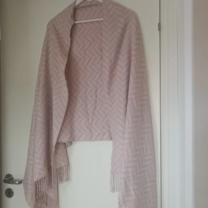 Rosa oversized sjal från Love & Lore - Säljer en superfin ljusrosa oversized sjal från Love & Lore. Den är mjuk och har ett subtilt mönster som ger den lite extra stil. Perfekt för att hålla sig varm under höst och vinter. Den har också fransar i ändarna som ger en snygg touch. Använd den för att piffa upp vilken outfit som helst!
