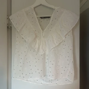 Vit blus med volangdetaljer från Zara - Säljer en superfin vit blus från Zara. Den har vackra volangdetaljer och ett broderat mönster. Blusen är ärmlös och har en luftig passform.