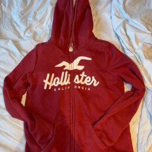 Röd hoodie från Hollister - Säljer en supermysig röd hoodie från Hollister med vit text och logga på framsidan. Den har en dragkedja och är fodrad med mjuk, vit fluff på insidan. Perfekt för kyliga dagar när du vill vara både varm och stilren. 🧥