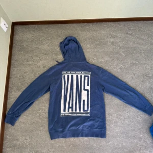 Blå hoodie från Vans - Säljer en snygg blå hoodie från Vans. Den har en stor logga på ryggen och en mindre logga på bröstet. Perfekt för en avslappnad stil och passar bra till både jeans och joggers. Den är i bra skick och superbekväm att ha på sig. Perfekt för höst och vår!