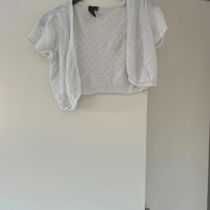 Vit kortärmad bolerotopp - Säljer en söt vit bolerotopp med korta ärmar. Den är stickad med ett fint hålmönster och har en luftig och lätt känsla. Perfekt för sommarens varma dagar eller som ett extra lager över en klänning. Passar både till vardags och fest!