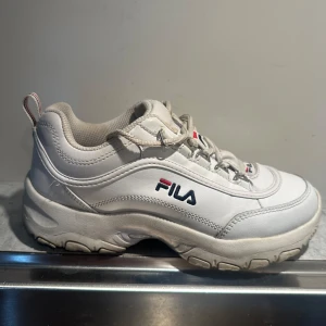 Vita sneakers från Fila - Säljer ett par vita Fila sneakers i bra skick. De har en chunky sula och klassisk snörning. Perfekta för både vardag och träning. Fila-loggan syns tydligt på sidan och tungan av skon.