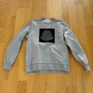 Moncler tröja - Säljer en snygg grå sweatshirt från Moncler. Den har ett svart och grått emblem på bröstet som verkligen sticker ut. Tröjan är långärmad och gjord i ett mjukt material som är perfekt för både höst och vinter. Pris kan diskuteras! Hör av er vid frågor