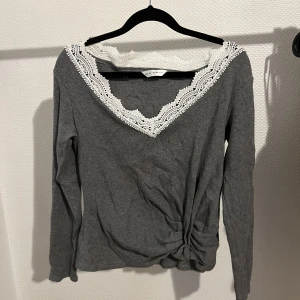 Grå tröja med spetsdetaljer - Säljer en superfin grå tröja med v-ringad spetsdetalj runt halsen. Tröjan har långa ärmar och en snygg knutdetalj framtill. Perfekt för både vardag och fest! 🖤