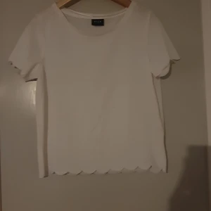 Vit topp med volangdetaljer - Säljer en söt vit topp från Vila. Den har korta ärmar och fina volangdetaljer vid ärmslut och nederkant. Perfekt för en somrig look eller en avslappnad dag ute. Toppen är i bra skick och redo för en ny ägare!